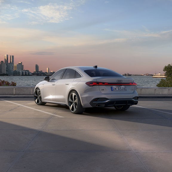 A sleek white car is parked on a waterside pavement at sunset, with a city skyline in the background and a ferry on the water to the right.
