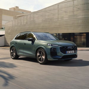 A green Audi Q3 drives past a modern building on a sunny day. The car's sleek design and motion evoke a sense of style and sophistication.