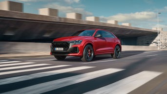 Der RS Q8 SUV performance schräg von vorn nähert sich einem Zebrastreifen. Rechts daneben ein futuristisches Gebäude in Betonoptik.