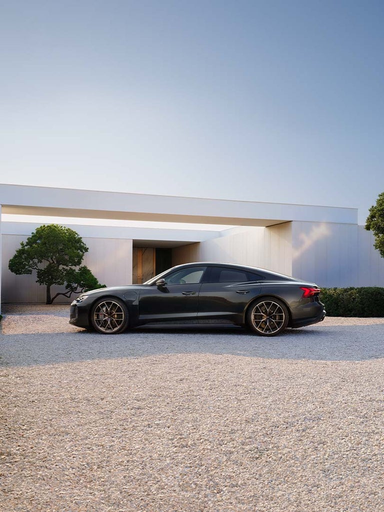 Dunkelgrauer Audi RS e-tron GT performance seitlich vor einem modernen Haus mit weißer Fassade und grünem Baum auf Kiesuntergrund.