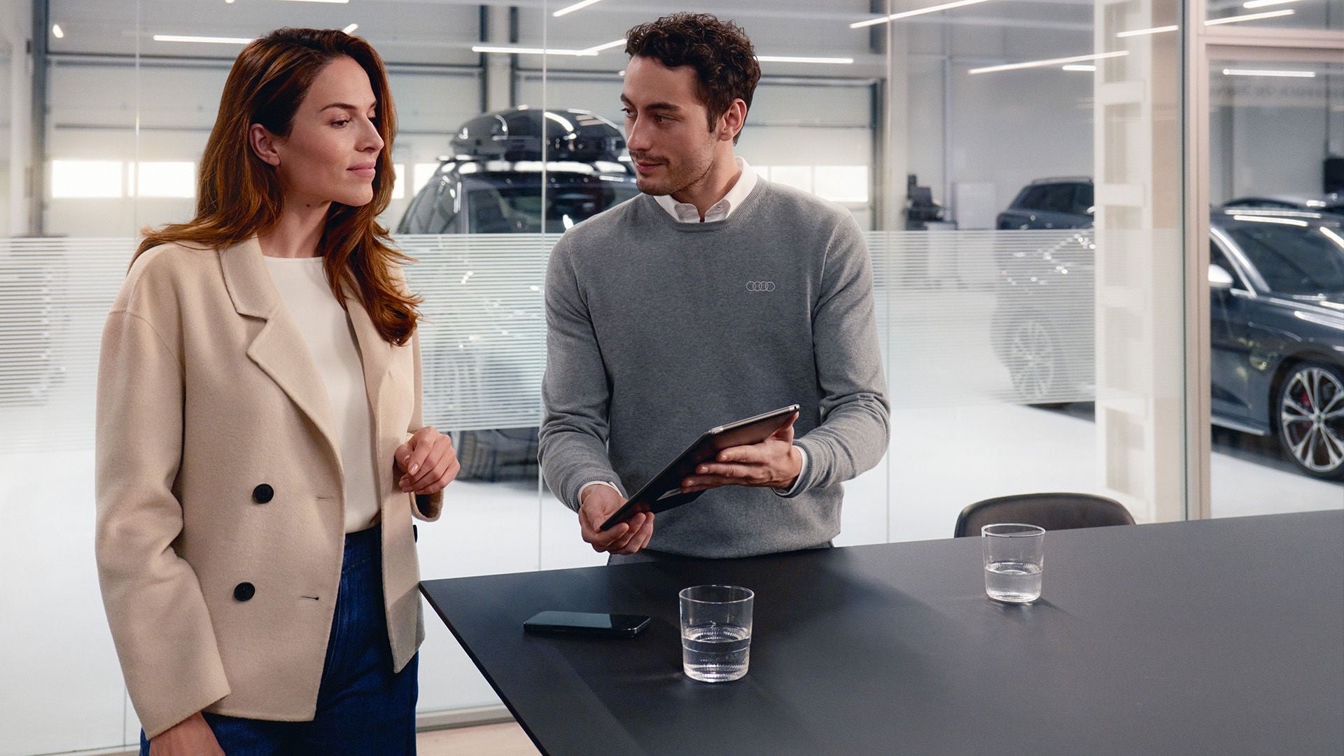 Eine Frau steht mit einem Audi Mitarbeiter an einem Tisch, der Audi Mitarbeiter hält ein Tablet in der Hand und zeigt der Frau etwas. Im Hintergrund sind Audi Modelle sichtbar.