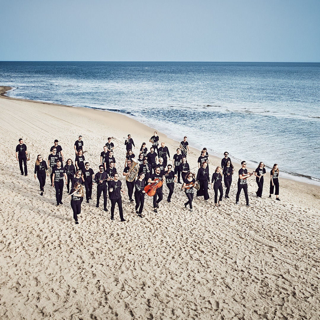 Orchester in schwarzen T-Shirts mit Instrumenten spaziert in einer Gruppe an einem Sandstrand entlang.