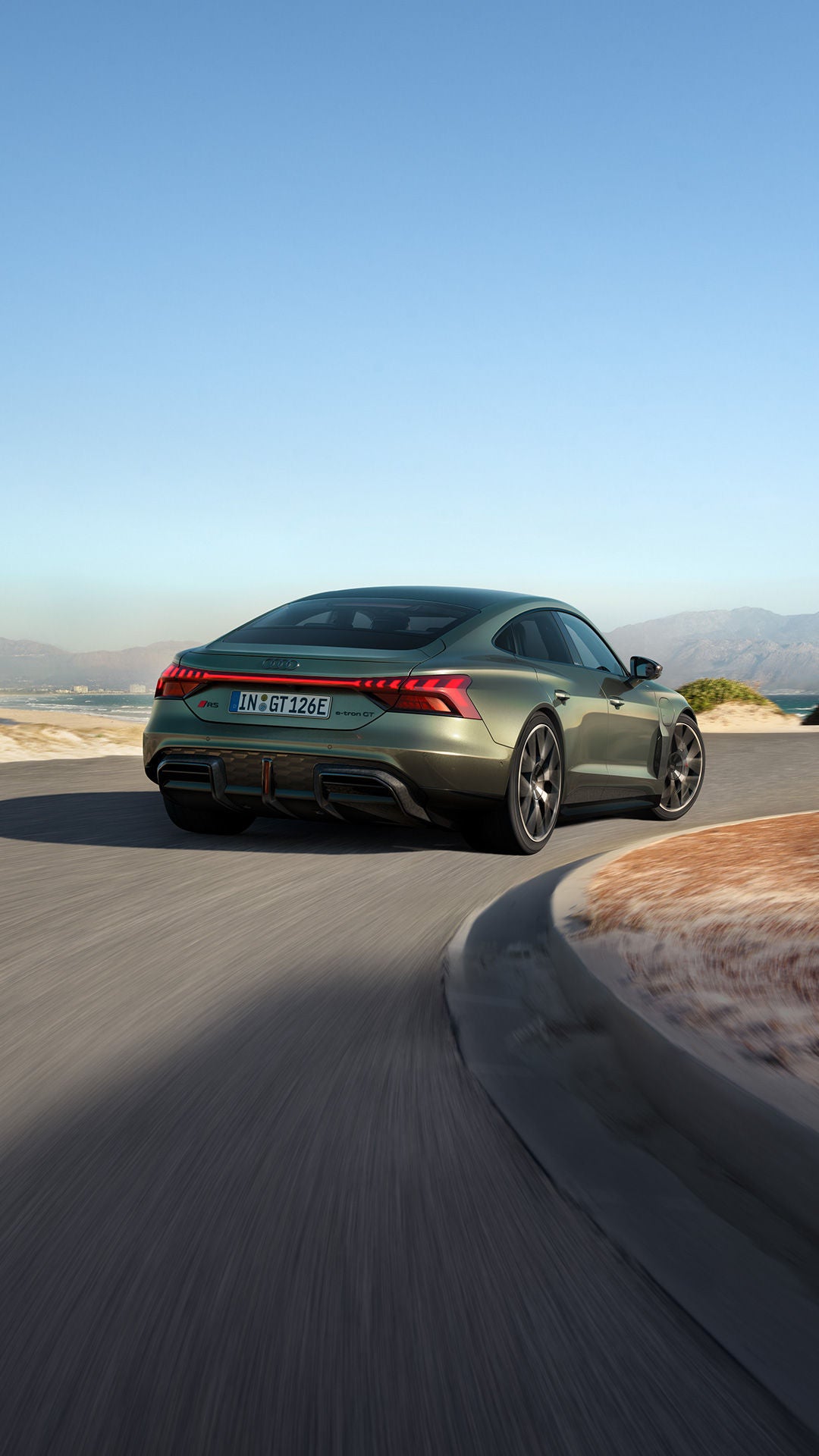 Ein grüner Audi RS e-tron GT performance fährt auf einer kurvigen Küstenstraße, mit Bergen, Meer und klarem Himmel im Hintergrund.