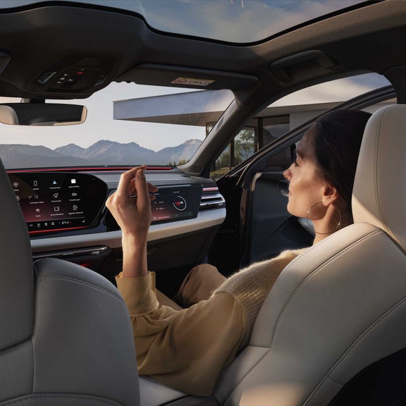 Dunkelhaarige Frau im beigen Oberteil sitzt entspannt im Audi A6 Avant, Blick auf Touchdisplay und Berge durch Panoramadach im Hintergrund.