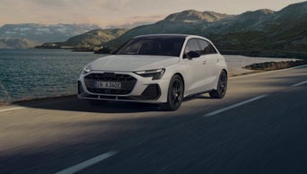 A silver Q7 SUV TFSI e is parked in front of a charging station, with a young, fashionable couple in the foreground.	