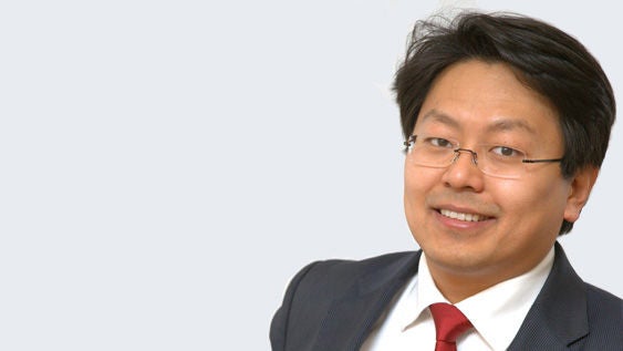 Chan-jo Jun: Porträt eines asiatischen, lächelnden Mann mit Brille im Anzug vor hellgrauem Hintergrund.