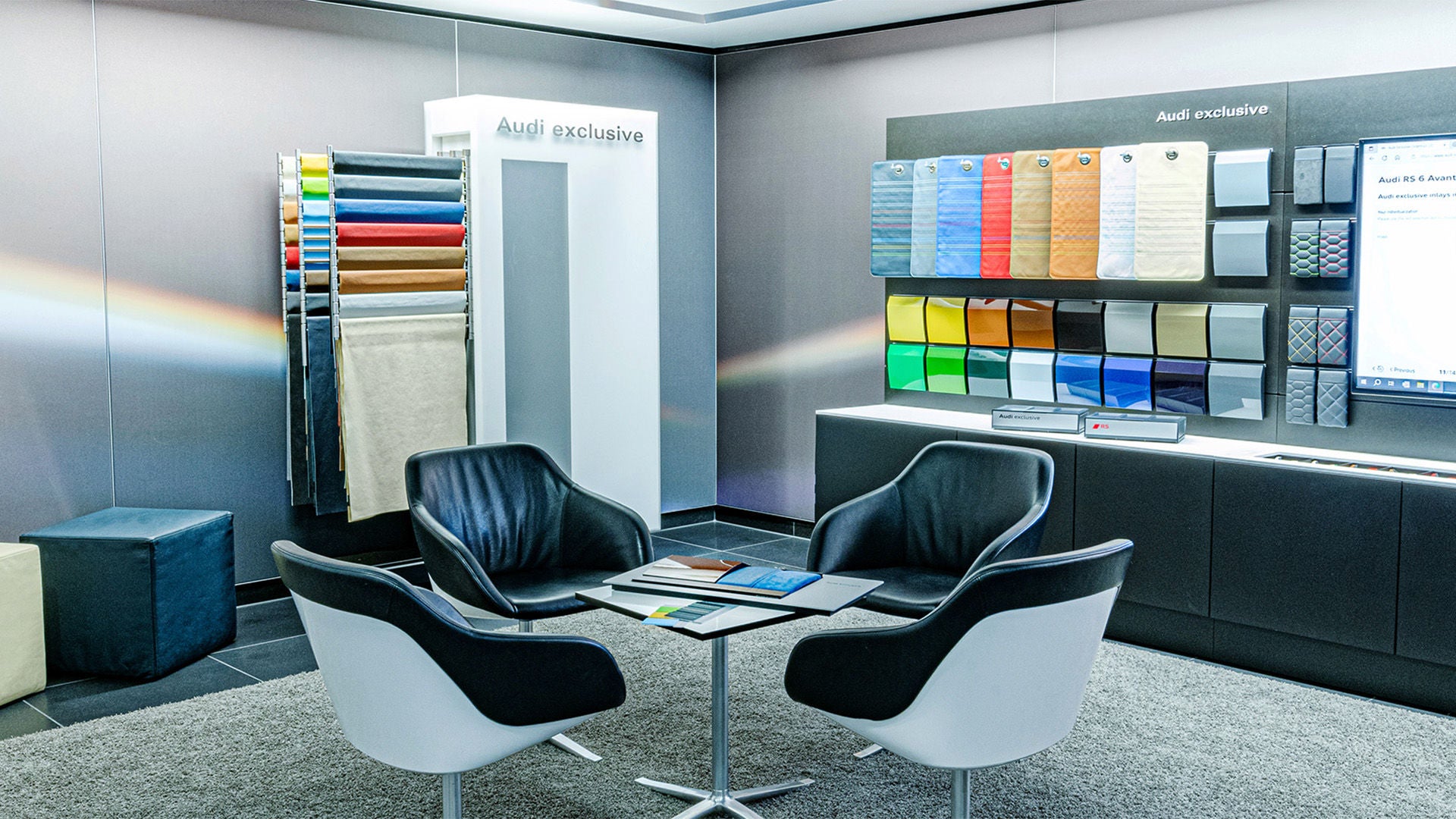 Room with fabric and color sample walls, four armchairs and a table with catalogs, labeled “Audi exclusive”.