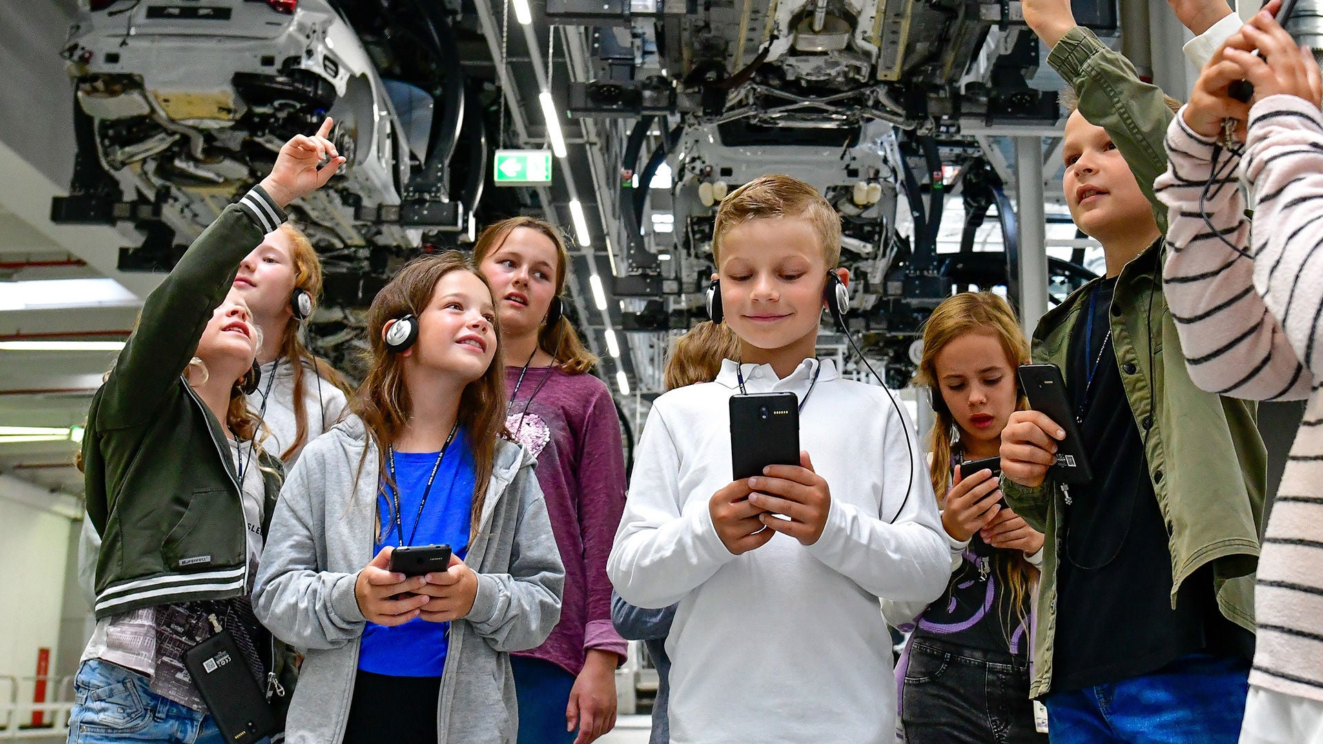 Gruppe von Kindern mit Kopfhörern bei einer Werksführung, einige zeigen auf Produktionsmaschinen.