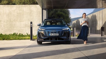 Frau steht neben einem dunkelblauen Audi vor einer modernen Garage im Freien, aufgenommen bei Tageslicht.