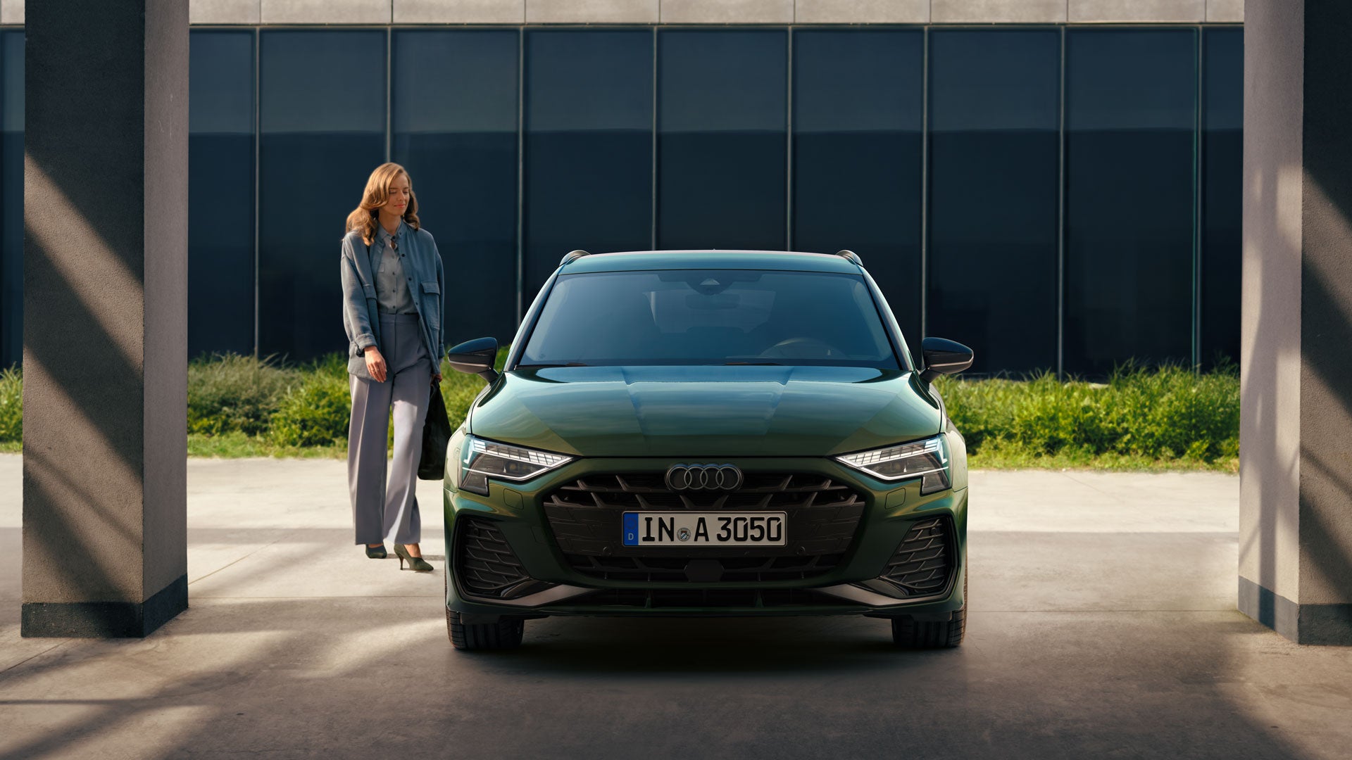 Eine Frau steht neben einem grünen Audi A3, frontal aufgenommen, vor einem modernen Gebäude mit dunklen Glasflächen.