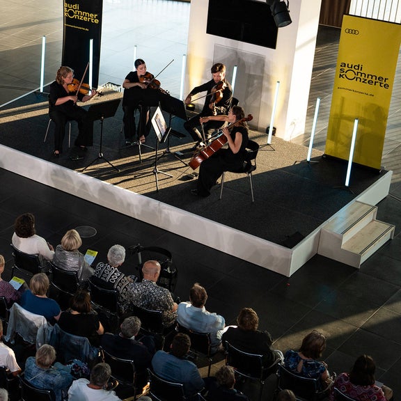 Streichquartett spielt auf einer kleinen Bühne vor sitzendem Publikum in einem hellen Foyer.