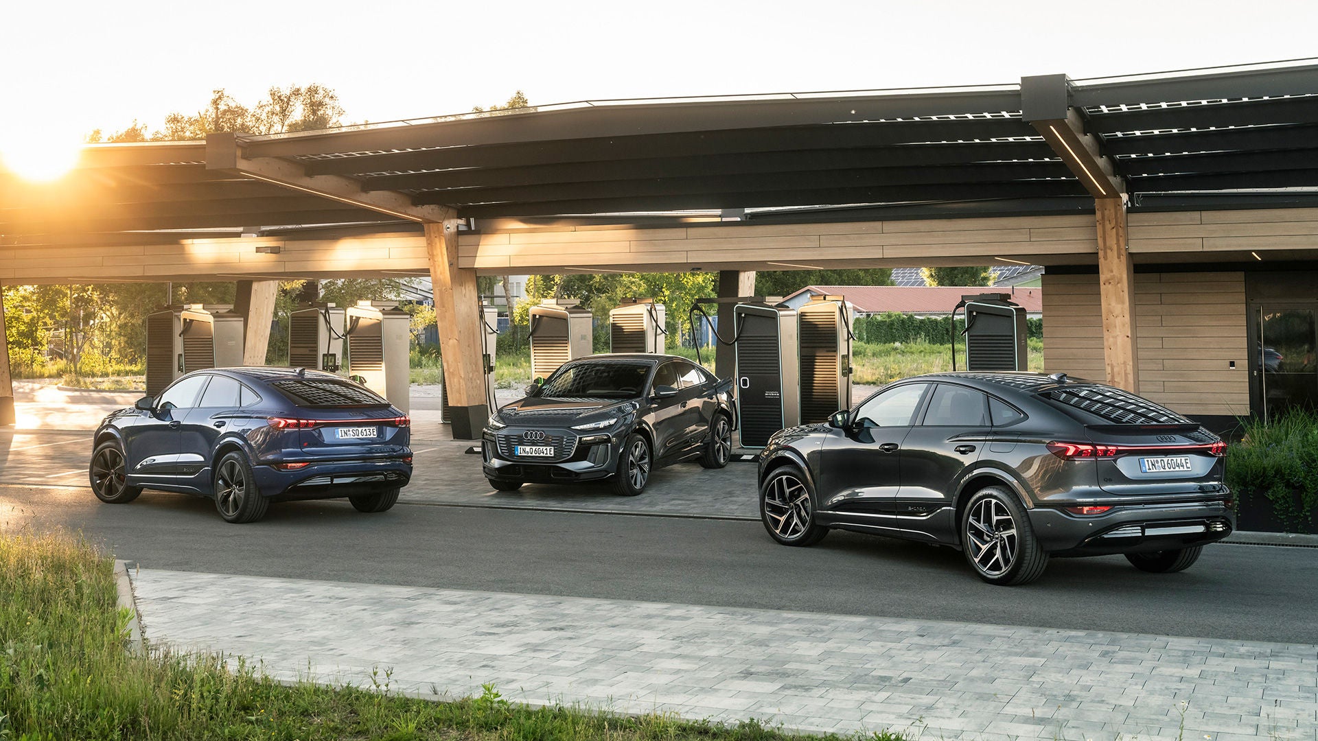 Three black Audi models are parked at a charging station. The middle Audi model is currently charging: the sun is setting in the background.