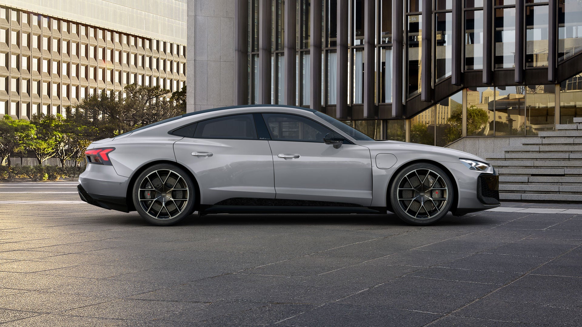 Grauer Audi RS e-tron GT performance geparkt vor einem modernen Gebäude mit Glasfassade und Treppe bei Sonnenuntergang.