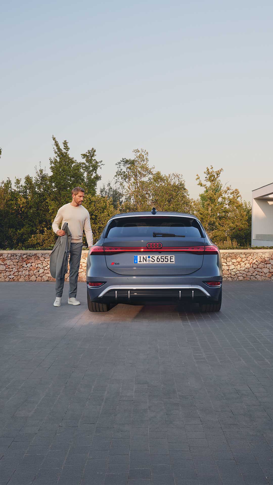Rückansicht eines grauen Audi. An der Fahrertür steht ein blonder Mann mit einer grauen Jacke in der Hand. Er scheint kurz davor, in das Auto einzusteigen.      