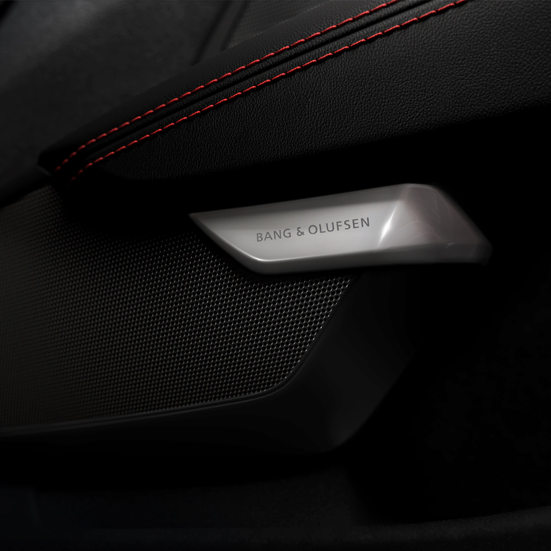 Black leather car interior with red stitching, silver Bang & Olufsen speaker grille, and black mesh panel.