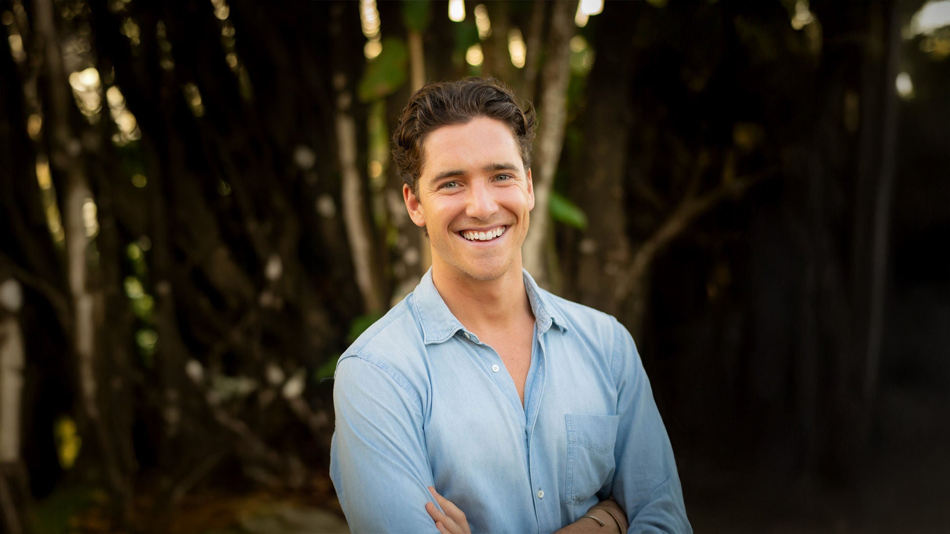 Mann im hellblauen Hemd, lächelnd mit verschränkten Armen, vor dunklem Hintergrund mit Bäumen.