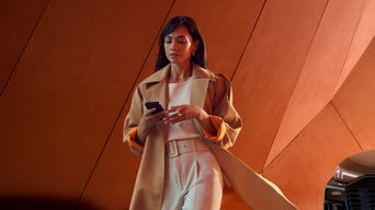 Frau im beigen Trenchcoat geht mit Smartphone vor orangefarbener Wand, rechts ist ein Audi-Frontscheinwerfer erkennbar.