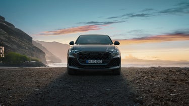 Der RS Q8 SUV von vorn an einem Strand auf Schotter, im Hintergrund das Meer und seitlich hervorragende Berge.