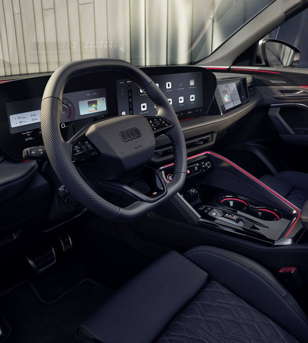 Een zicht op de cockpit van de Audi SQ5 Sportback door de zijruit aan bestuurderszijde. De verschillende displays in de cockpit zijn zichtbaar, net als de rode sfeerverlichting.