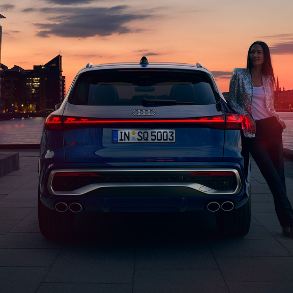 Blue Audi SQ5 parked by riverside at sunset with city skyline in background.