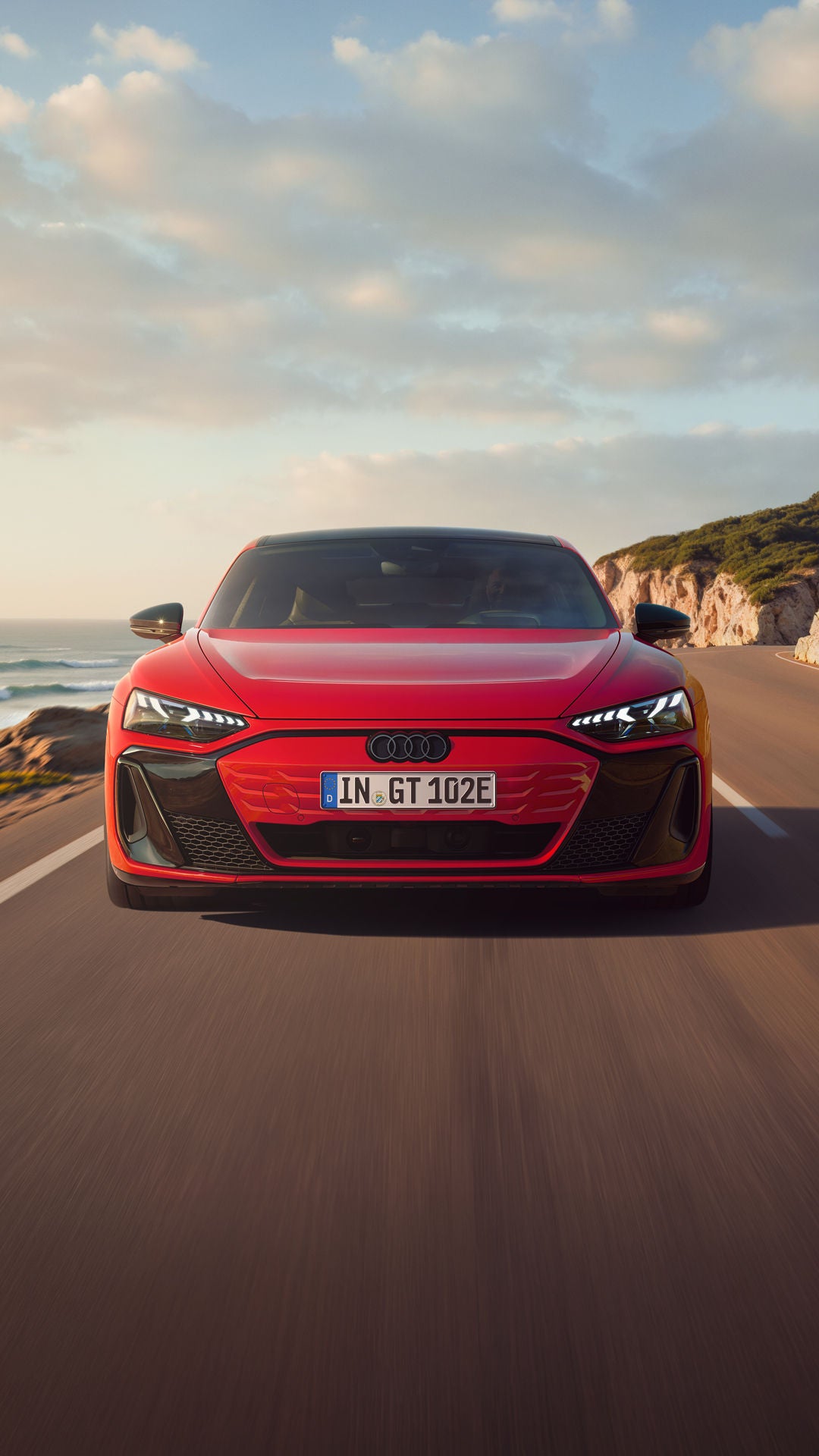 Frontansicht eines roten Audi e-tron GT quattro, der auf einer Küstenstraße fährt. Im Hintergrund sind Klippen, das Meer und der Himmel zu erkennen. Die gesamte Szenerie ist in das Licht eines Sonnenuntergangs gehüllt.
