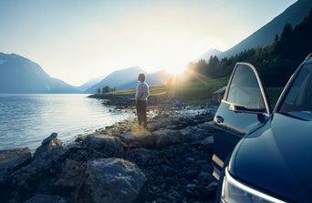 A person stands by a tranquil lake at sunrise, with mountains in the background and an open car door nearby.