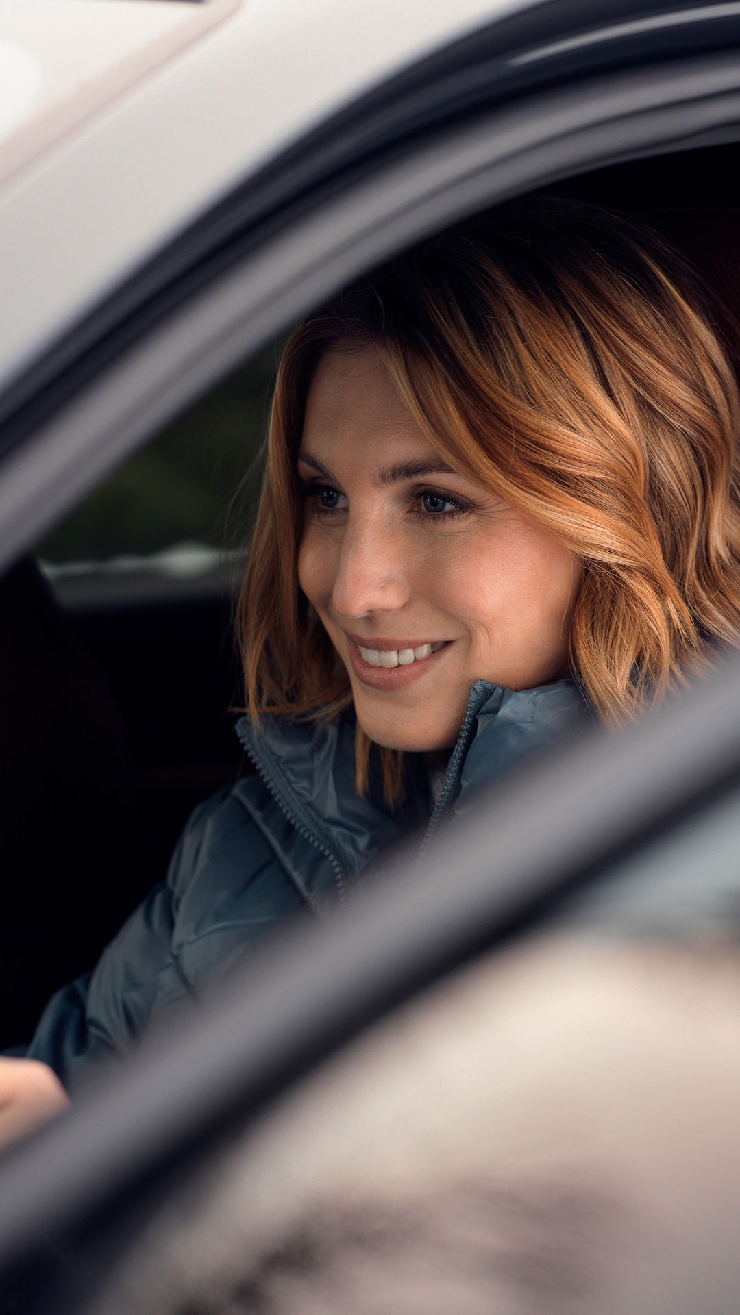 Frau mit blondem Haar und blauer Jacke sitzt konzentriert am Steuer eines Fahrzeugs.