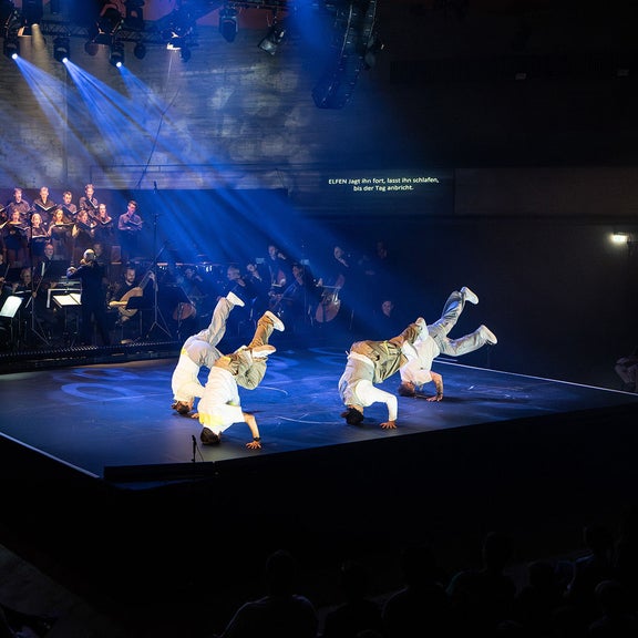 Vier Tänzer in weißer Kleidung bewegen sich auf der Bühne vor Orchester und Chor, blaues Bühnenlicht.