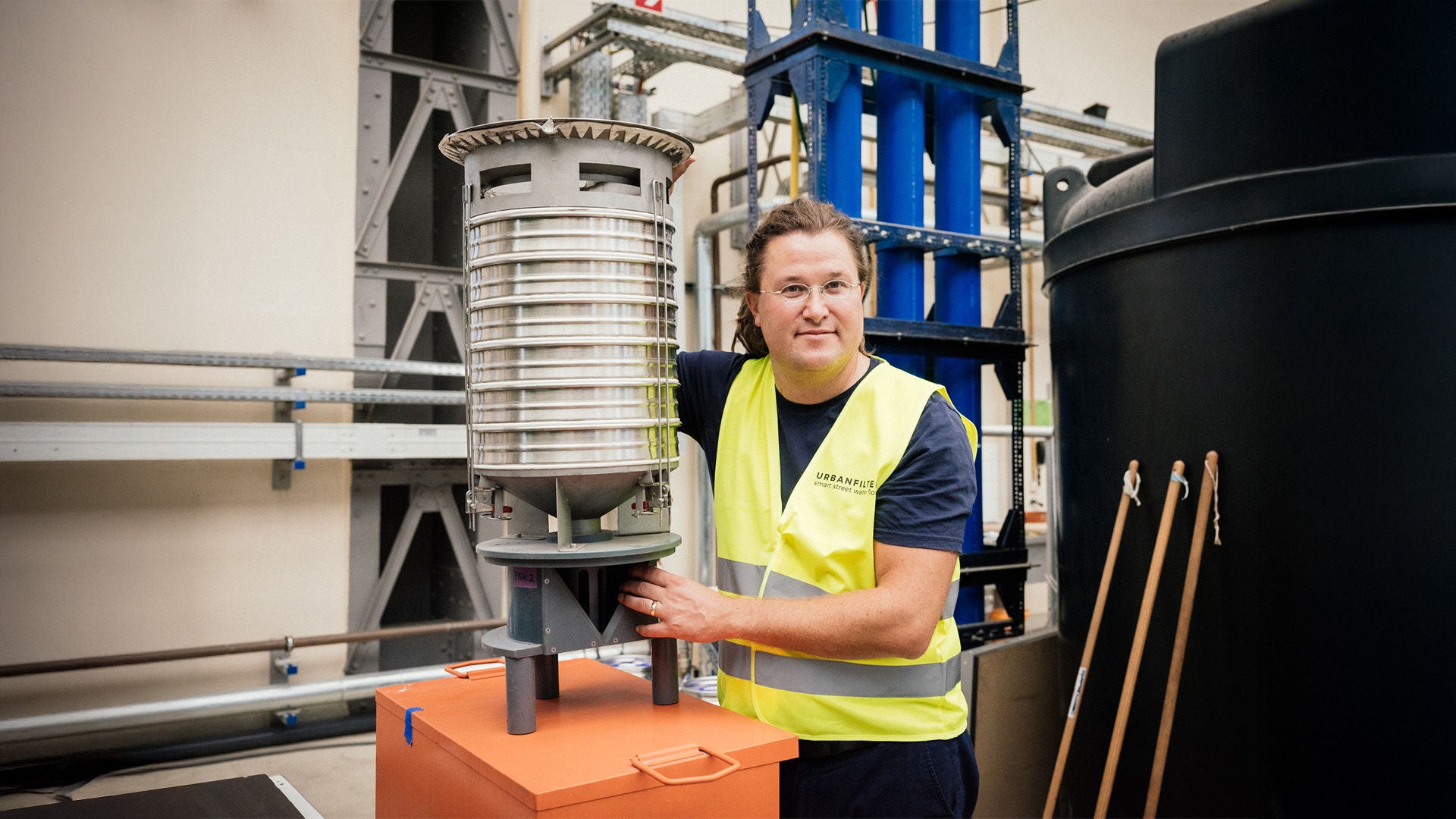 Man wearing a safety vest stands in a workshop presenting a cylindrical filter system on an orange box.