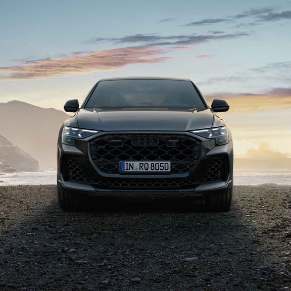 Ein grauer Audi RS Q8 SUV von vorn vor Küstenlandschaft bei Sonnenuntergang auf steinigem Boden.