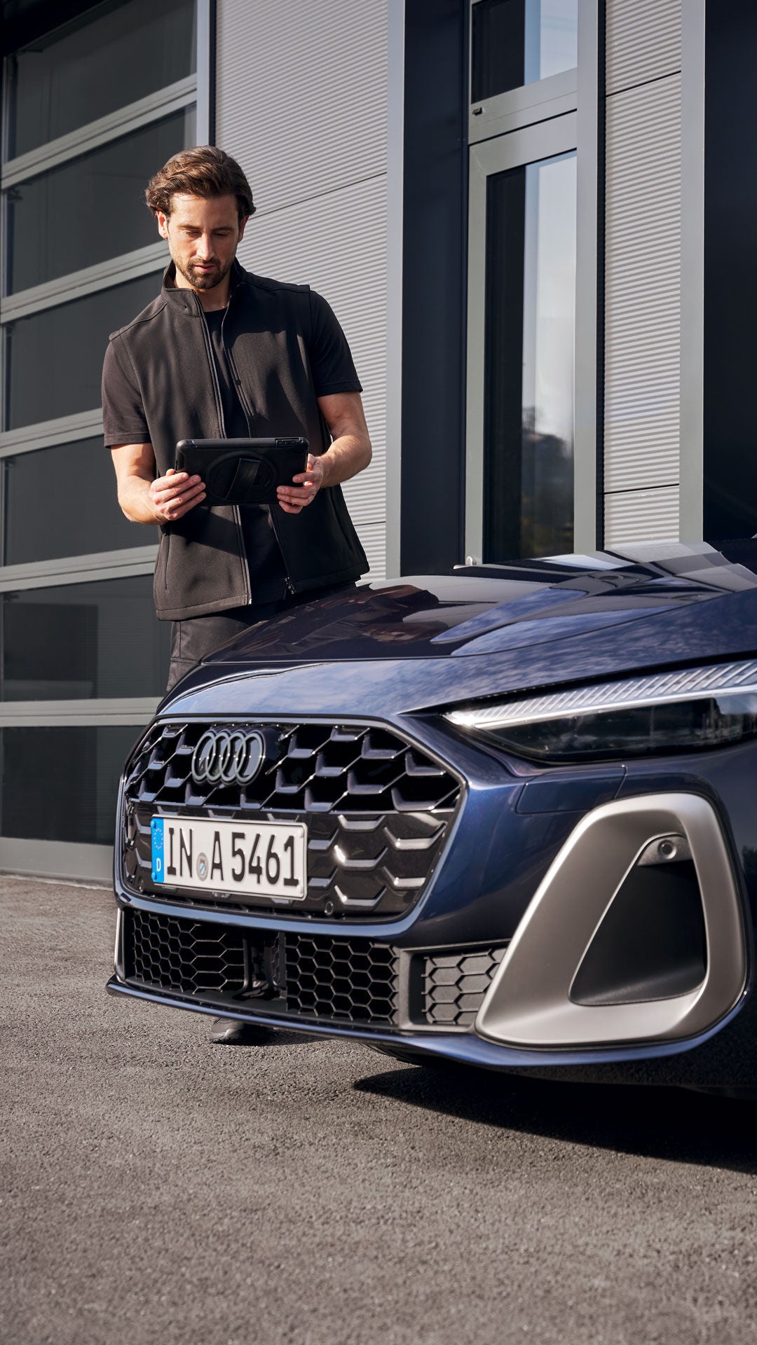 Mann steht vor einem blauen Audi und bedient ein Tablet, vor moderner Gebäudefassade mit Glas- und Metallflächen.