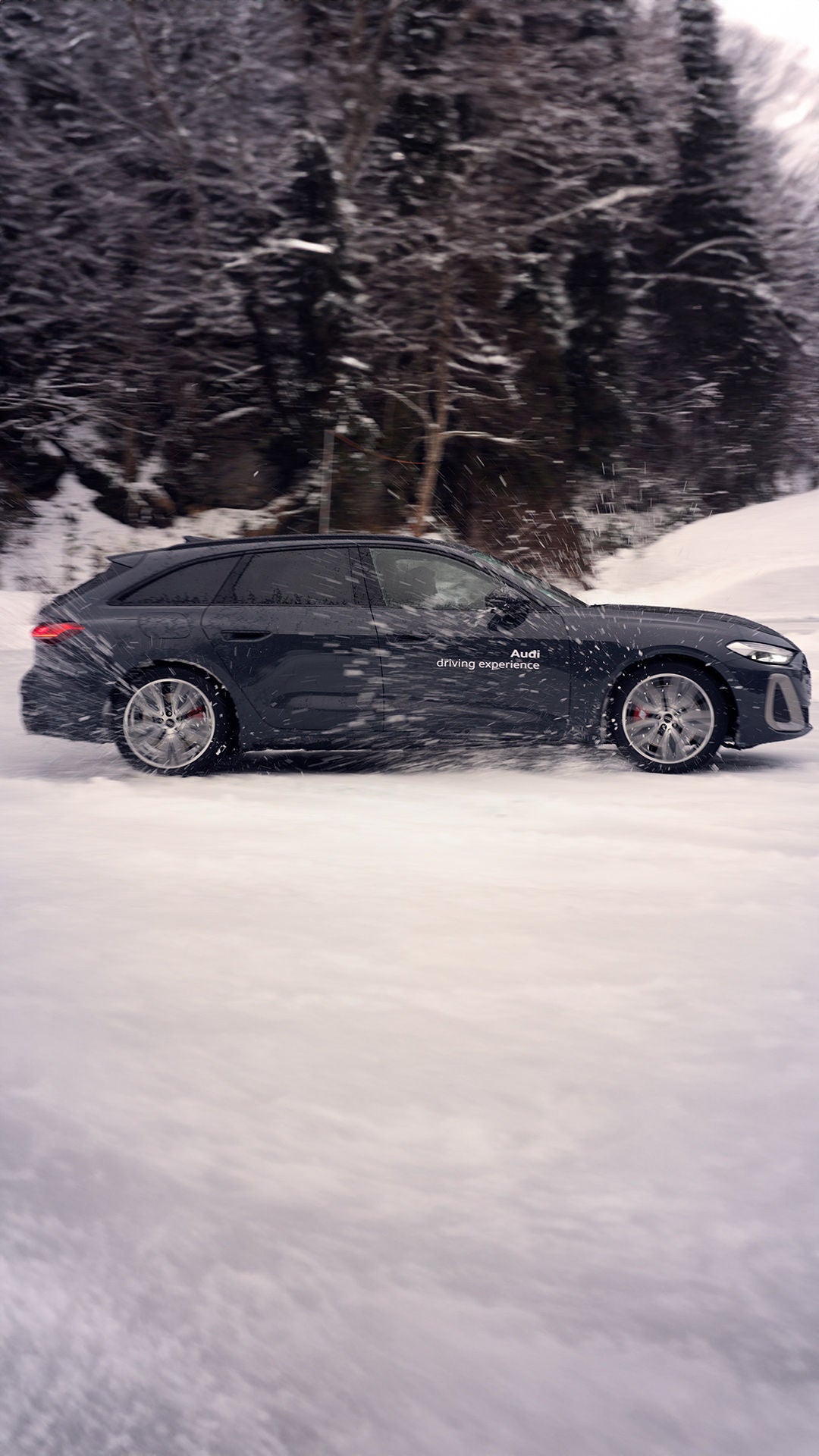 Dunkler Audi S5 Avant mit weißem Schriftzug „Audi driving experience“ fährt auf schneebedeckter Straße, aufgewirbelter Schnee im Fokus.