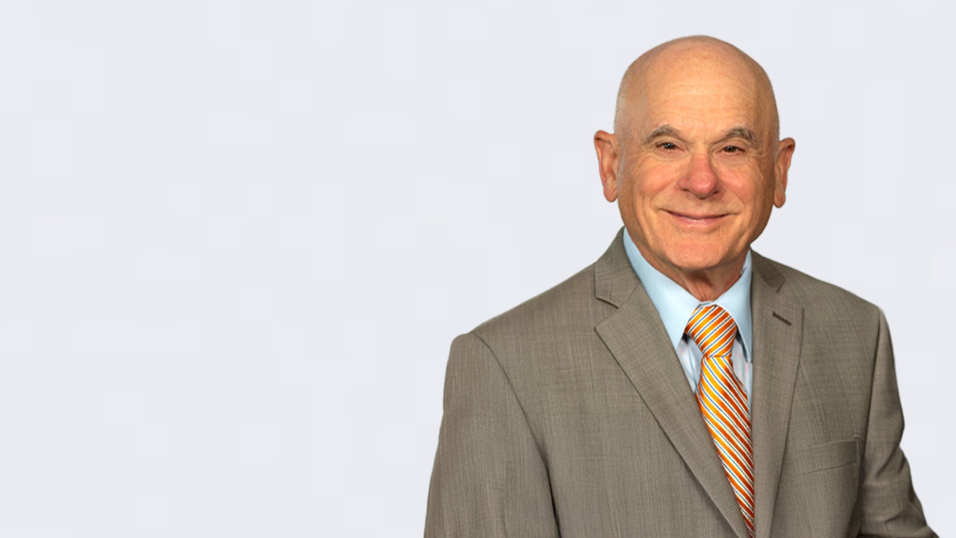 Mann im grauen Anzug mit blauem Shirt und gestreifter Krawatte steht vor einem hellgrauen Hintergrund.