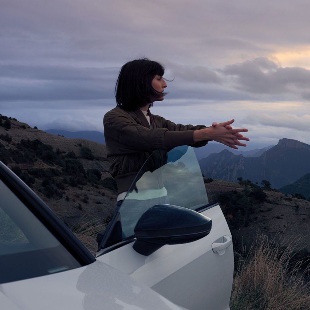 A person leans out of a white car against a backdrop of mountains and a cloudy sky, enjoying the scenic view.
