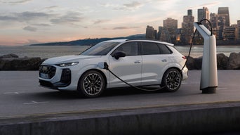 White Audi Q3 SUV e-hybrid plugged into a charging station beside a waterfront, with a city skyline at sunset in the background.
