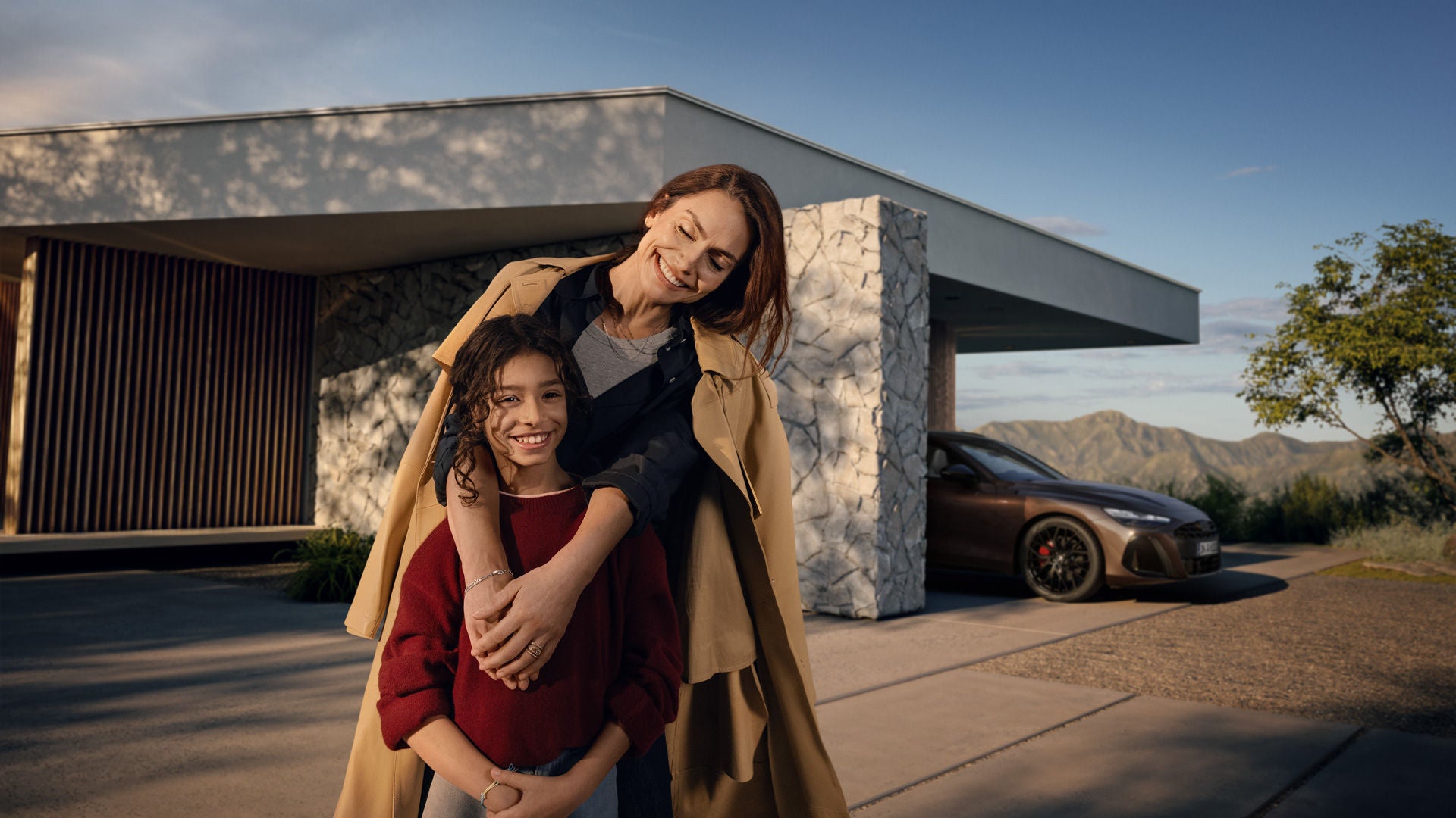 Eine Frau steht vor einer Garage und hat die Arme über ein lächelndes Kind gelegt. In der Garage ist ein brauner Audi A6 Avant geparkt.