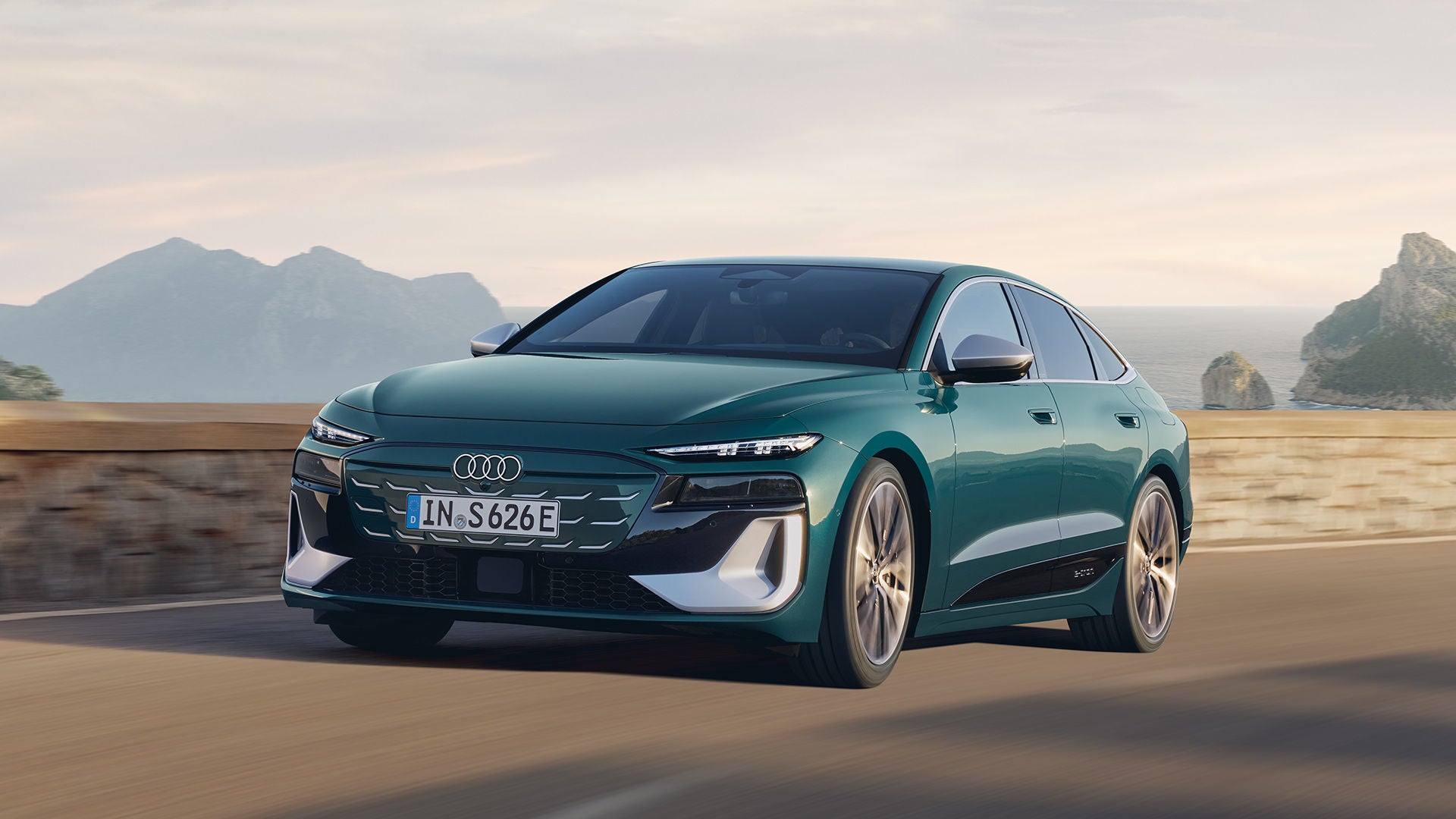 Dunkelgrüner Audi S6 Sportback e-tron fährt auf einer Küstenstraße vor bergiger Landschaft bei Tageslicht.