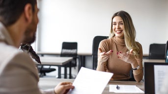 Job interview: A man and a younger woman are talking, the man can be seen from behind, holding documents in his hand and the woman is smiling