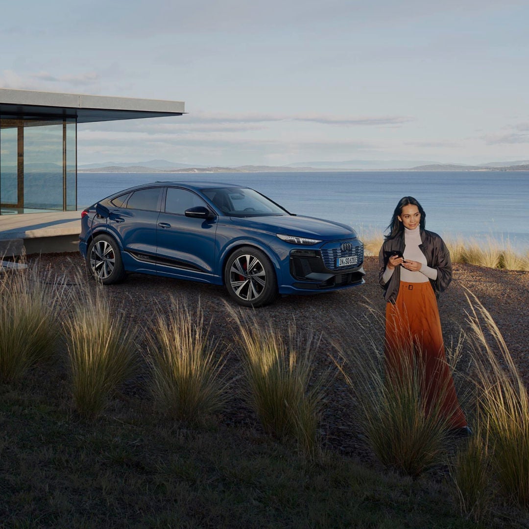 Woman stands on a grassy hill by the ocean, holding a phone with a blue SUV parked behind her