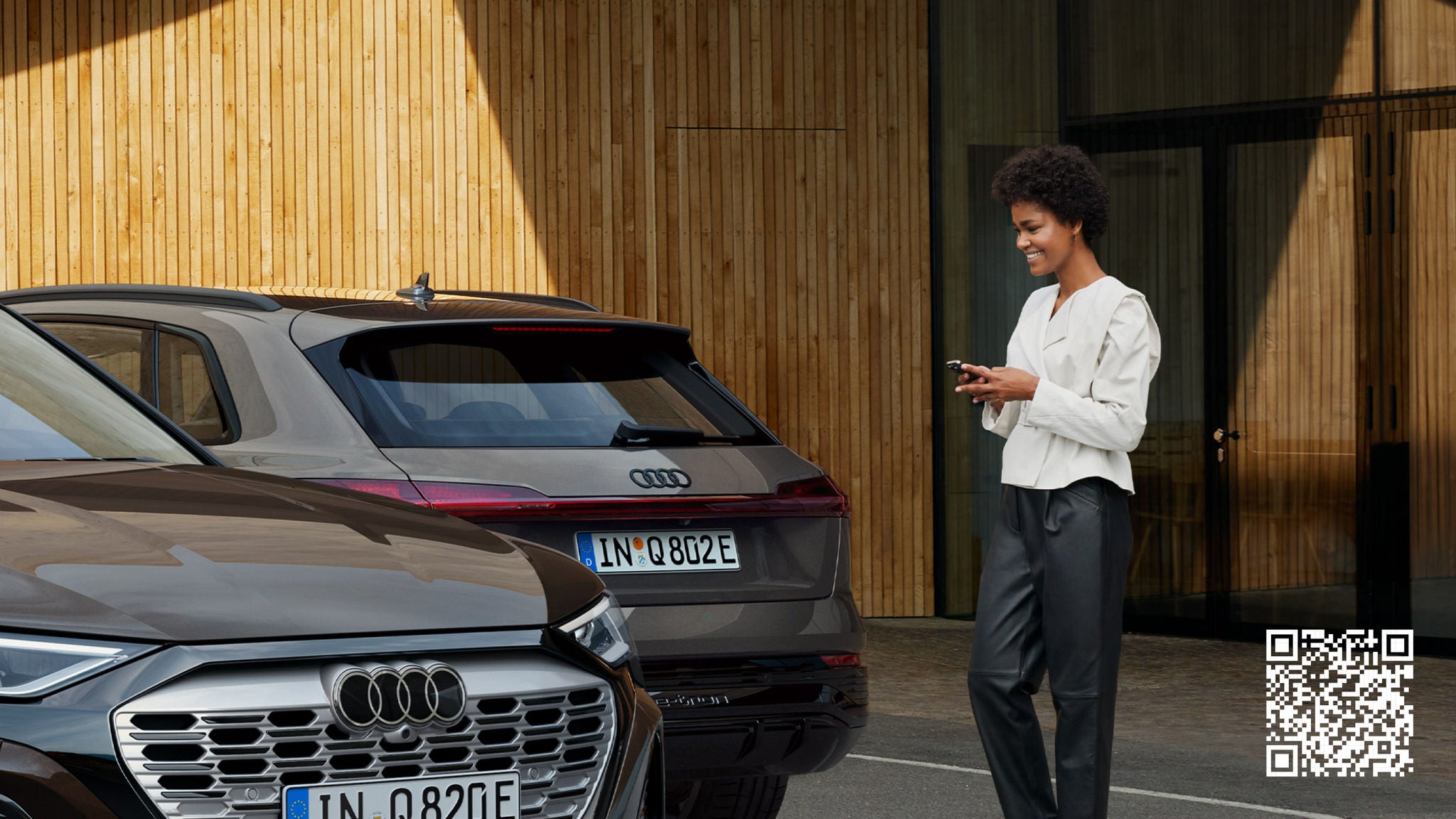 Zwei Audi-Fahrzeuge stehen nebeneinander, und eine Frau steht neben ihnen und schaut auf ihr Telefon.