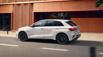 White hatchback car in motion on an urban road with modern buildings.