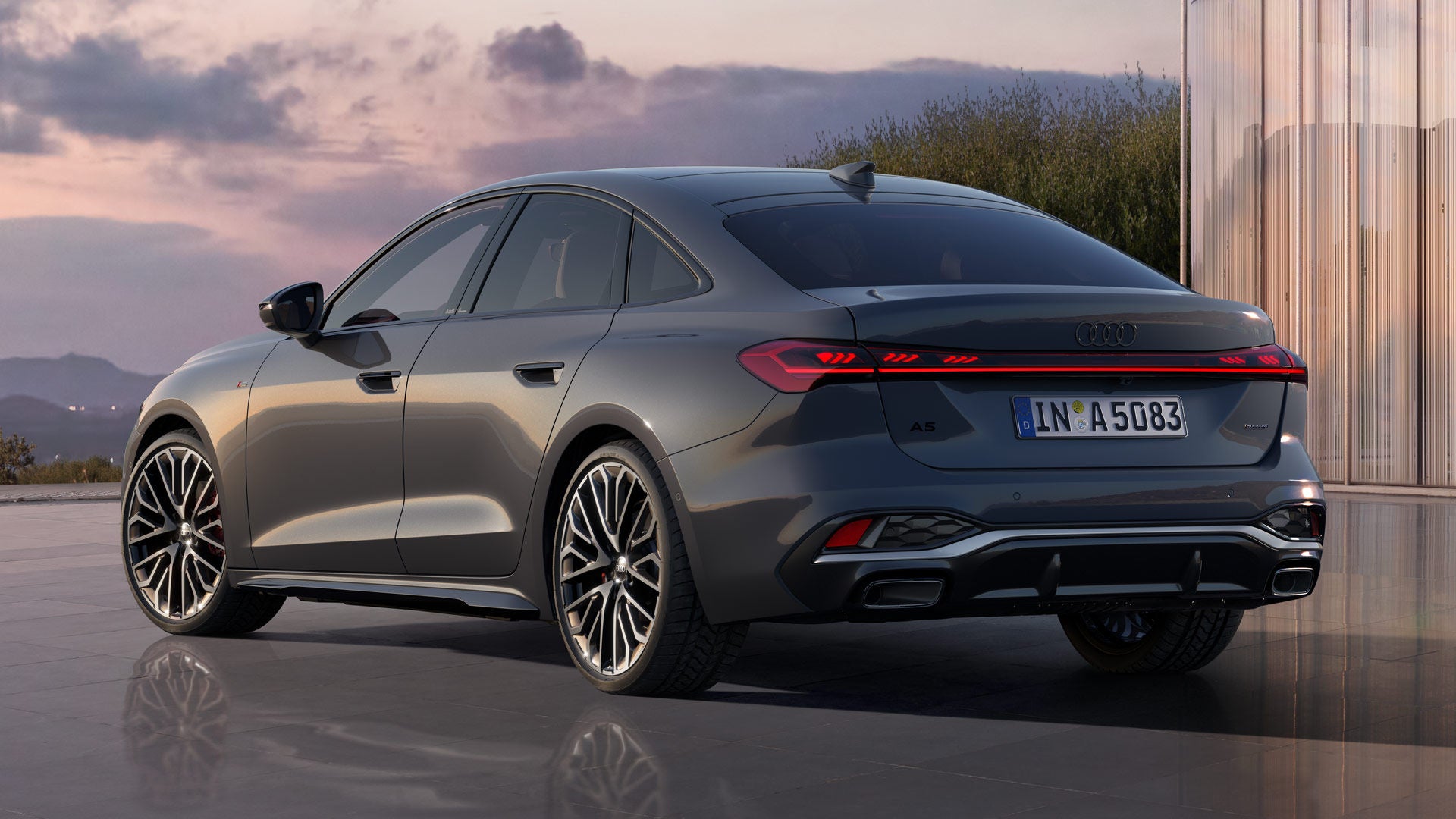 Eine graue Audi A5 Limousine steht vor einer Glasfassade bei Sonnenuntergang, Heck und Seitenlinie im Fokus.