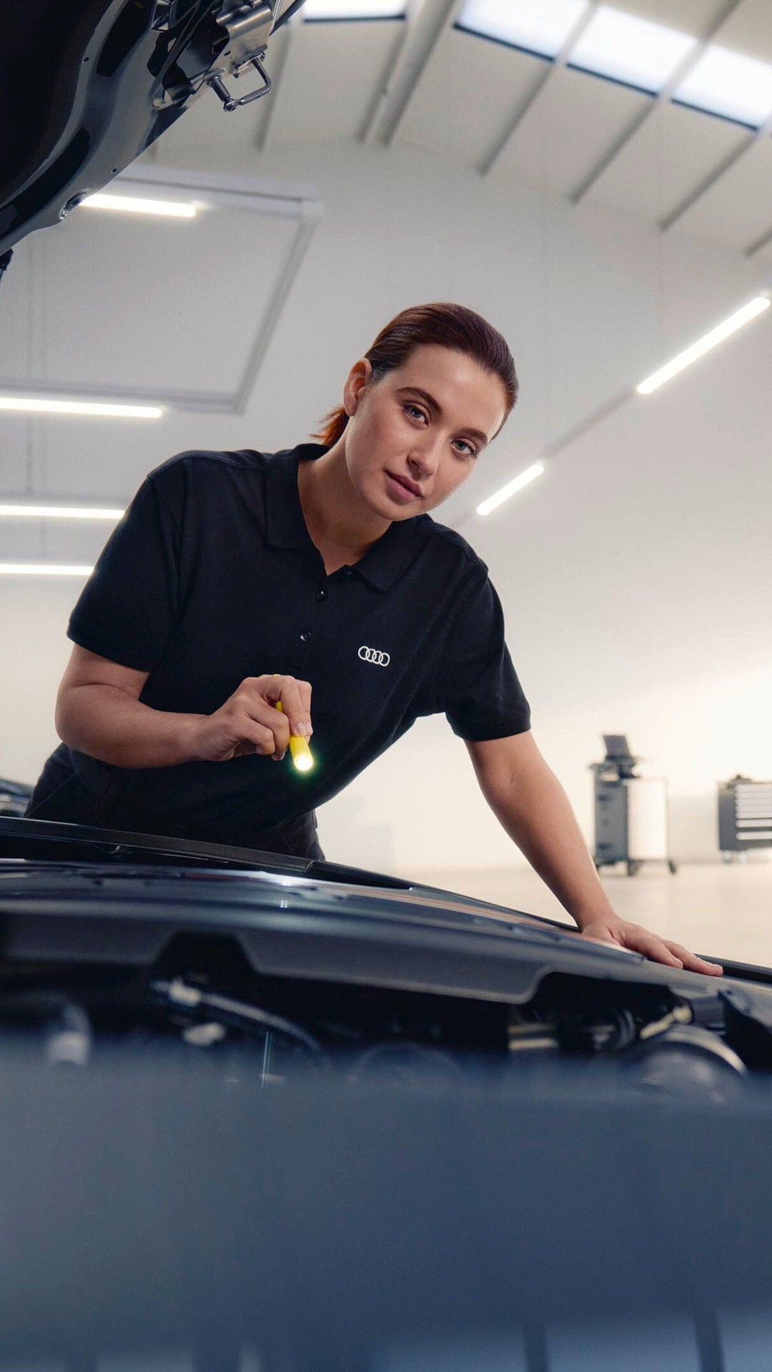 Mitarbeiterin prüft den Motorraum eines Autos mit einer Taschenlampe.