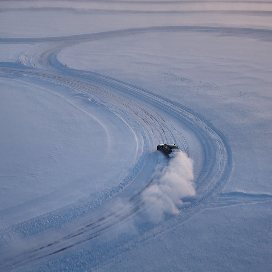 A dark car drives along a snow-covered circular road, kicking up a cloud of snow; wide tire tracks in the snow.