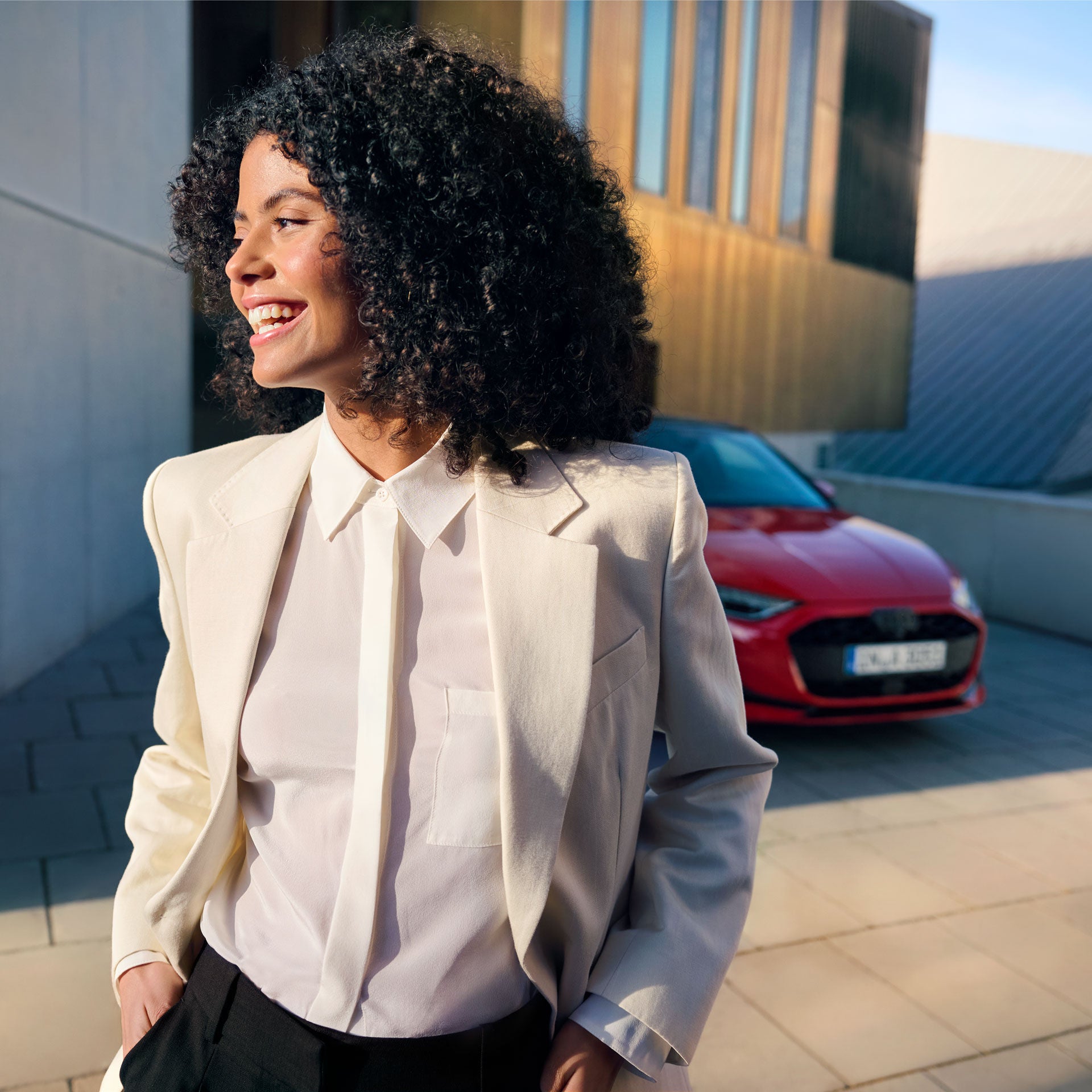 Eine lachende Frau im weißen Blazer steht im Sonnenlicht vor einem roten Auto und einem modernen Gebäude.