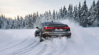 A dark green Audi RS 5 sedan drives along a snow-covered road; snow dust, a pine forest, and a blue sky. 