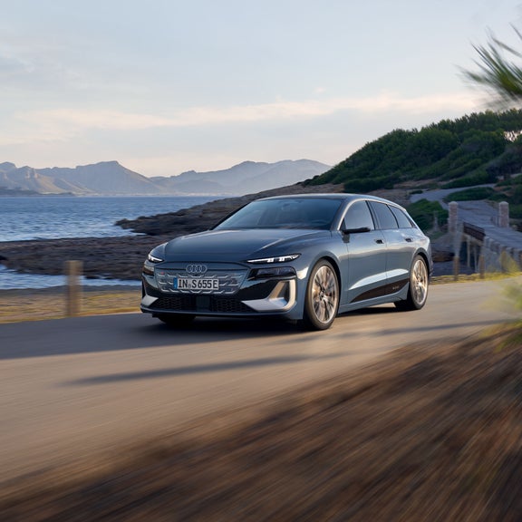 Der S6 Avant e-tron aus schräger Frontansicht, wie er eine Straße an der Küste entlangfährt. Im Hintergrund sind Berge zu sehen.
