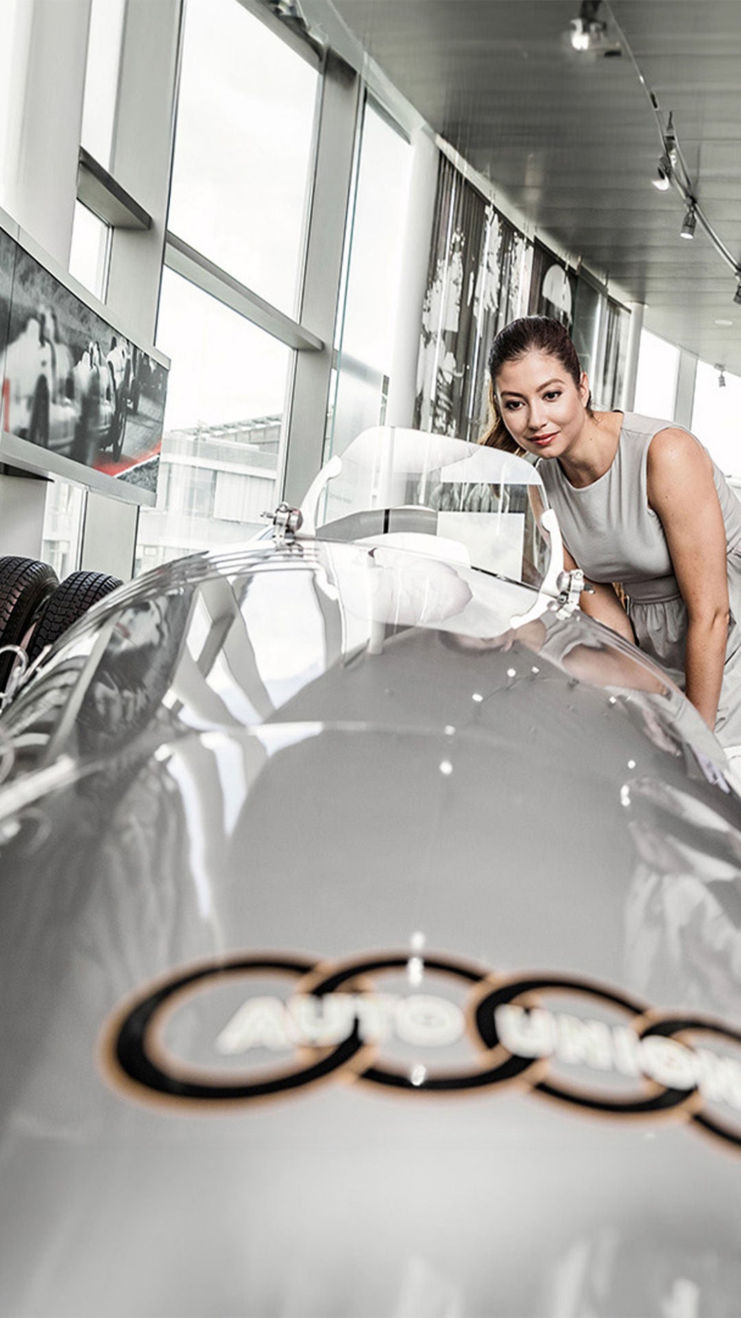 Three people look at a silver historic racing car with the Auto Union logo in an exhibition.