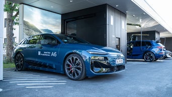 Drei blaue Audis stehen angeschlossen an Ladesäulen an einem Audi Charging Hub. Der A6 ist im Vordergrund und hat auf der Seite "Jetzt hier Probefahren" stehen.