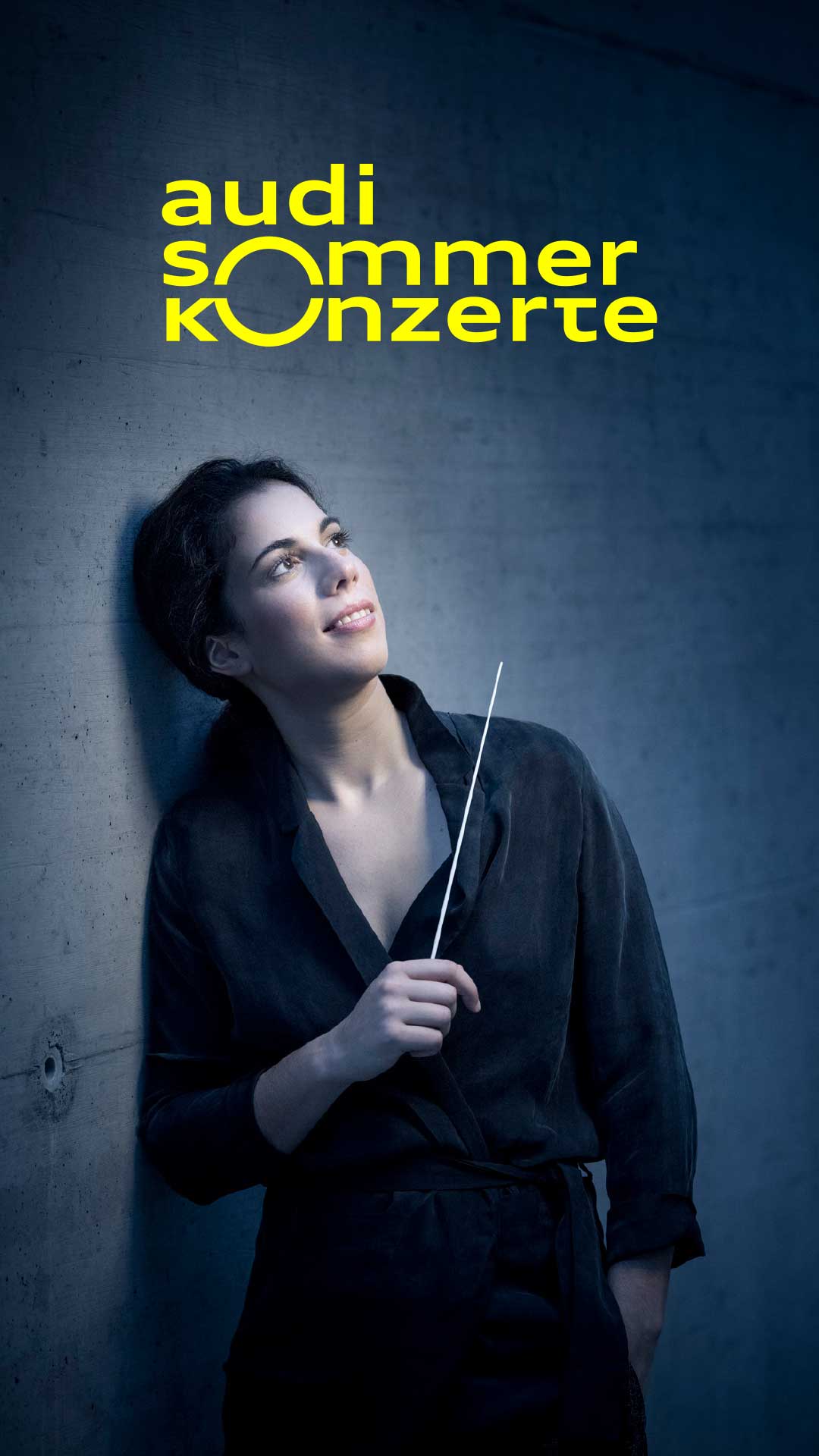 Frau mit Dirigentenstab lehnt an einer Wand, schaut nach oben; daneben Logo der Audi Sommerkonzerte in Gelb.